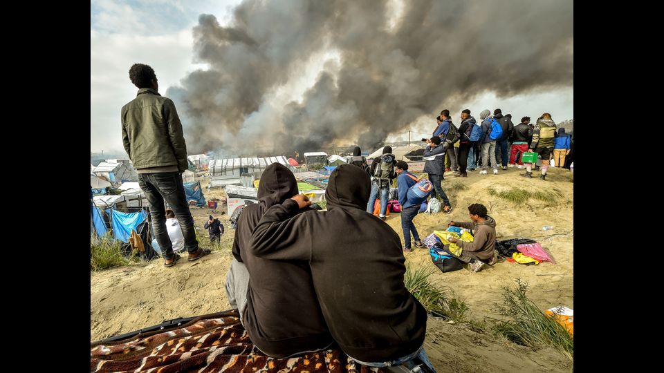 I migranti guardano il fumo degli incendi dal campo a Calais, nel nord della Francia, nel corso di una massiccia operazione per cancellare l'insediamento dove 6000-8000 persone hanno vissuto in condizioni disastrose (26 ottobre 2016) (foto Afp) &nbsp;