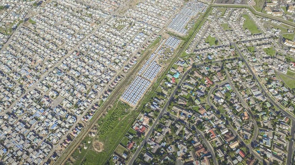 Johnny Miller &egrave; un antropologo che ha deciso di darsi alla fotografia per documentare le diseguaglianze. Il suo lavoro si &egrave; svolto principalmente in Sudafrica, ma anche Kenya e Messico sono state mete delle sue ricerche. Sul suo sito &nbsp;si trovano testimonianze di come in alcune metropoli del mondo ricchezza e povert&agrave; convivano fianco a fianco.Nomzamo/Lwandle &egrave; un 'hotspot' dei problemi economici, sociali e razziali del Sudafrica.   In origine pensato come luogo di residenza per lavoratori di colore durante l&rsquo;apartheid, conta oggi 60.000 abitanti. Intorno vi sono state costruite le comunit&agrave; di Strand e di Somerset West. La prima in particolare &egrave; una cittadina piuttosto benestante. Nella foto, Strand sulla destra e Somerset West sulla sinistra: in mezzo, gli accampamenti di Nomzano/Lwandle, circondati da ringhiere. Questo nucleo abitativo &egrave; stato sgomberato dalle autorit&agrave;, ma successivamente le baracche sono state ricostruite.