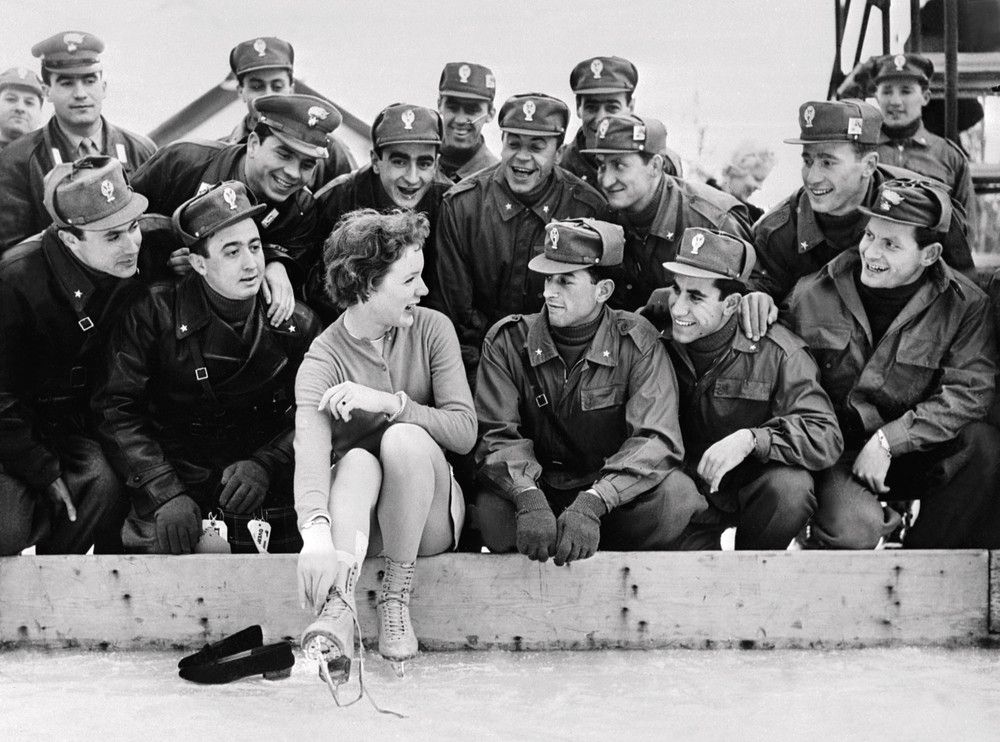 &nbsp;La pattinatrice statunitense Tenley Albright seduta sul bordo della pista di pattinaggio con un gruppo di soldati italiani durante la cerimonia di apertura delle Olimpiadi invernali del 1956 a Cortina