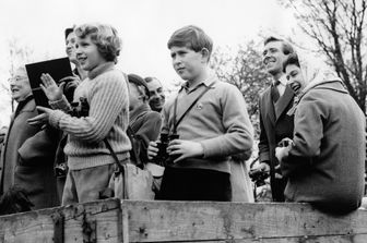 La regina Elisabetta, con il capo coperto da un foulard, assiste a una gara campestre nel 1961 a Badminton. In primo piano, col binocolo, il principe Carlo