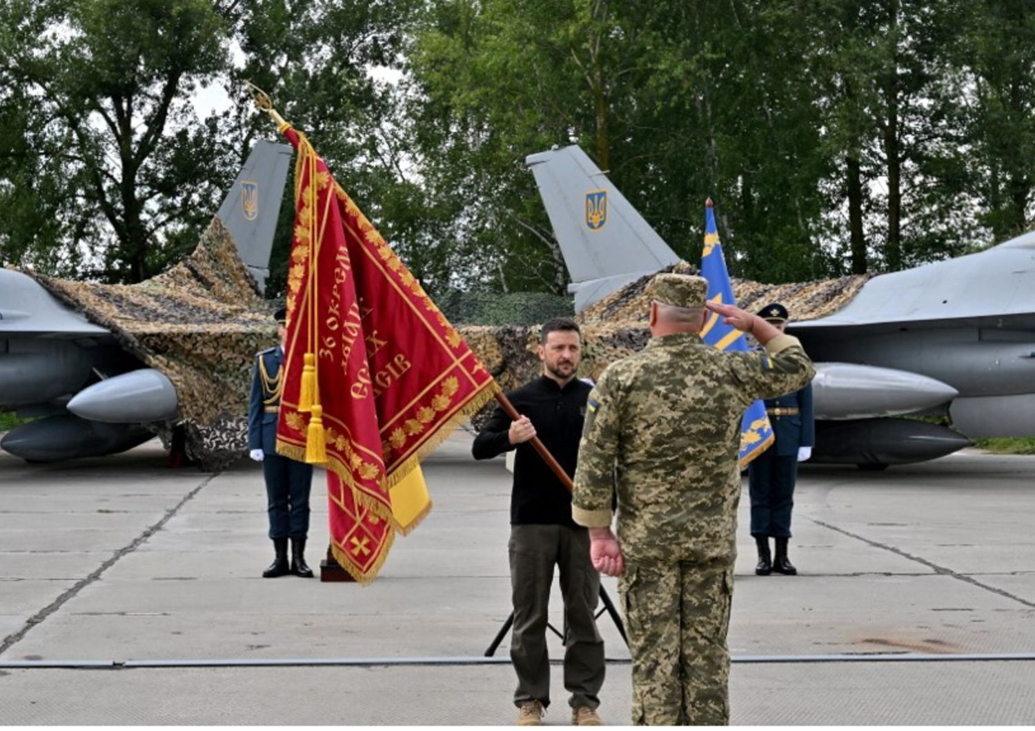 Il presidente ucraino Zelensky consegna le insegne di una bandiera a un militare mentre si trova di fronte a una coppia di caccia F16 durante una cerimonia tenuta in occasione della Giornata delle forze aeree ucraine il 4 agosto 2024