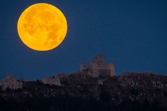 La luna piena di luglio, nota come "Luna del Cervo", tramonta dietro il castello di Rocca Calascio, vista da Castel del Monte (Abruzzo), Italia