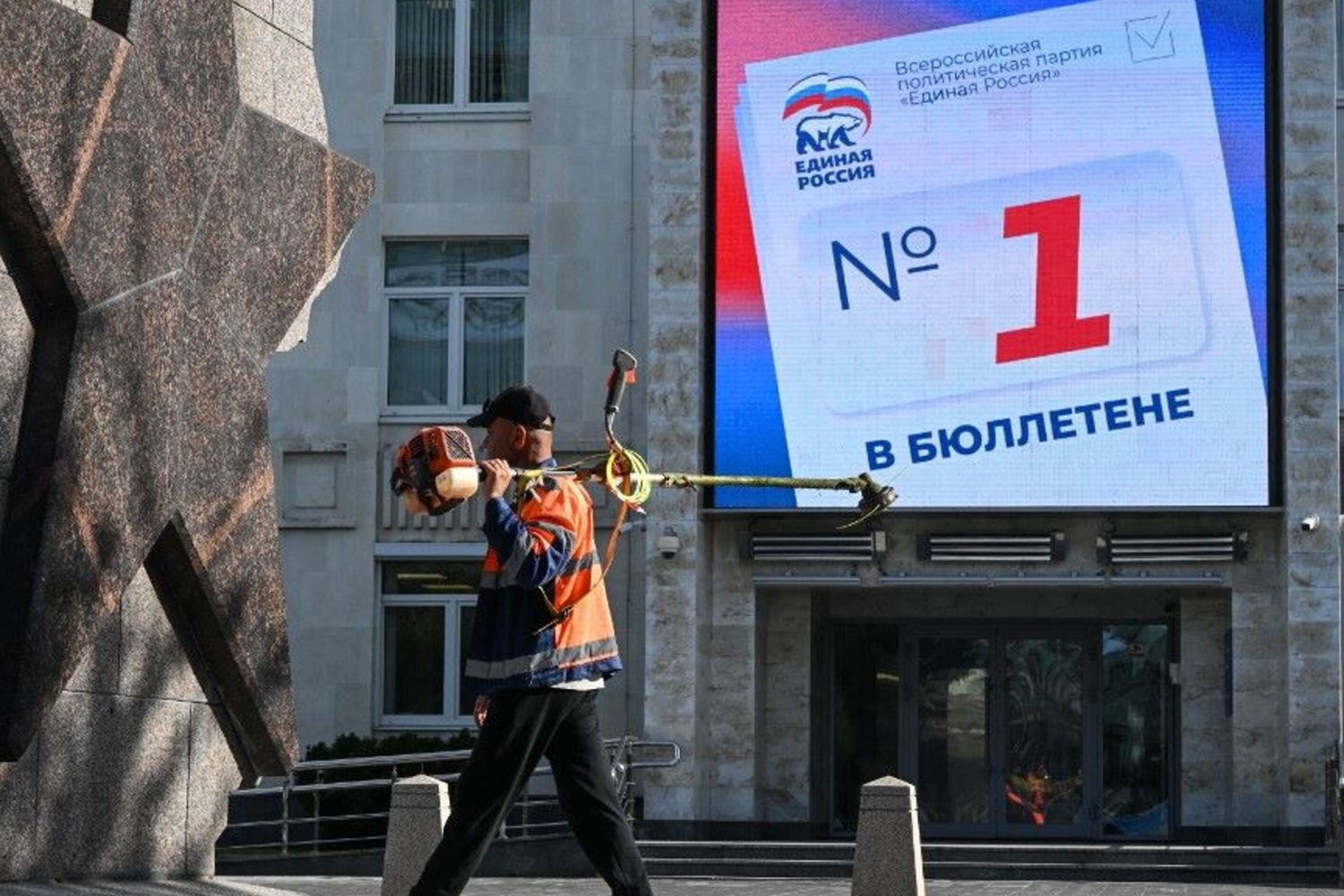 Un uomo con un tosaerba passa davanti a uno schermo digitale sull'edificio della sede del partito Russia Unita, che invita i cittadini a votare per i candidati del partito alle prossime elezioni per la Duma di Stato